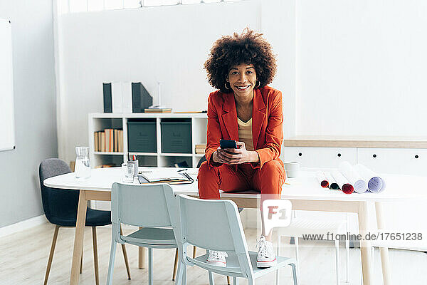 Smiling businesswoman with smart phone sitting on desk in office