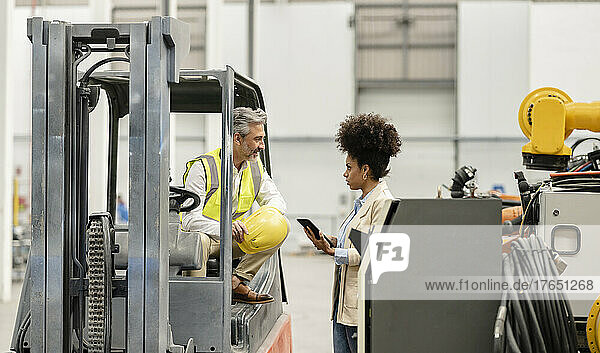 Geschäftsfrau hält Tablet-PC in der Hand und diskutiert mit Techniker  der im Gabelstapler in der Fabrik sitzt