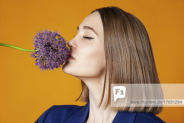Woman with eyes closed smelling allium flower against orange background