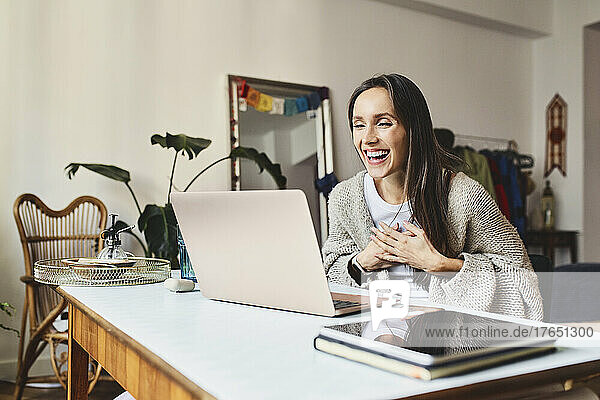 Laughing freelancer doing video call through laptop sitting at desk