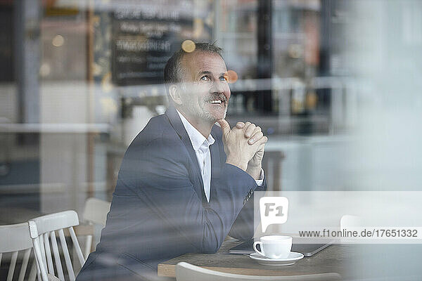 Smiling businessman with coffee cup and laptop seen through glass window at cafe