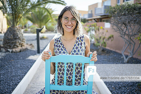 Glückliche junge Frau mit Brille sitzt auf einem blauen Stuhl auf dem Fußweg