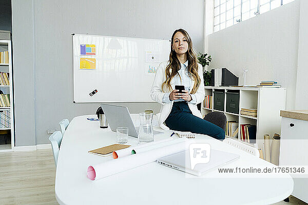 Businesswoman holding smart phone sitting on desk in office