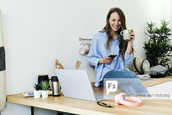 Smiling freelancer sitting on kitchen counter with mobile phone at home