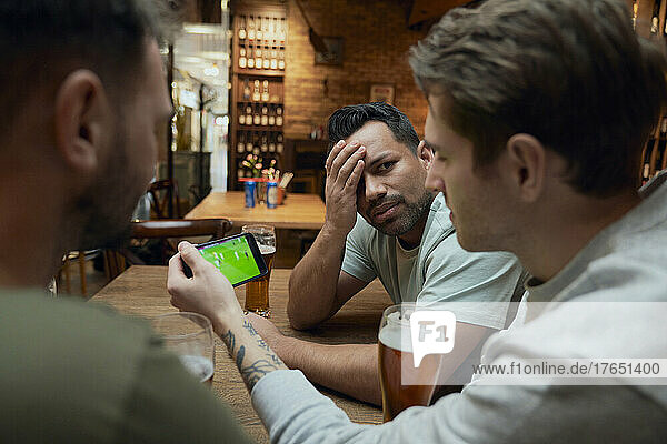 Drei frustrierte männliche Fußballfans trinken Bier und schauen sich ein Spiel auf dem Smartphone in einer Kneipe an