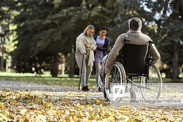 Senior disabled man sitting in wheelchair in front of nurse walking with woman at park