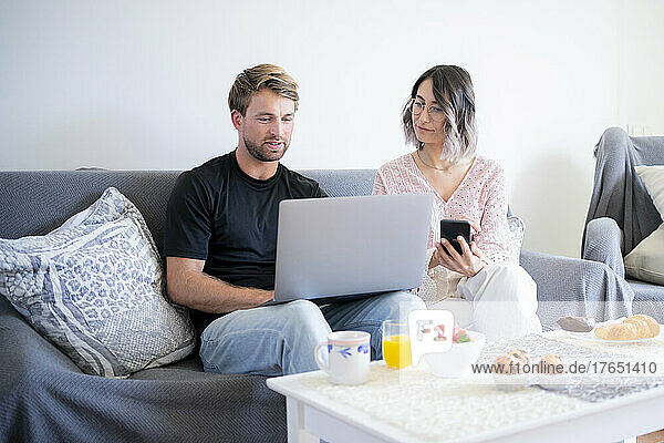 Man using laptop sitting by woman holding smart phone on sofa at home