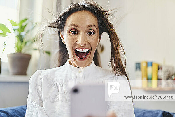 Überraschte Frau mit zerzausten Haaren  die zu Hause ihr Smartphone benutzt