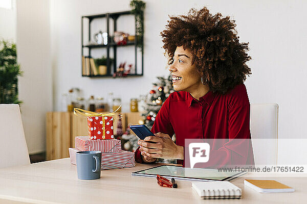Happy young woman with mobile phone sitting at table in living room