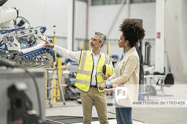 Engineer pointing at robotic arm discussing with businesswoman holding file folder in factory