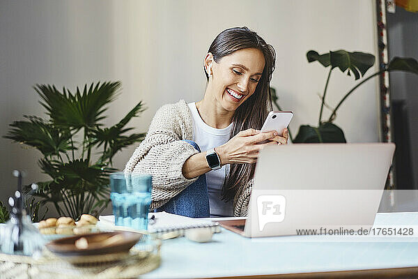 Happy freelancer using mobile phone sitting with laptop at desk