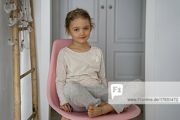 Cute girl sitting cross-legged on pink chair at home