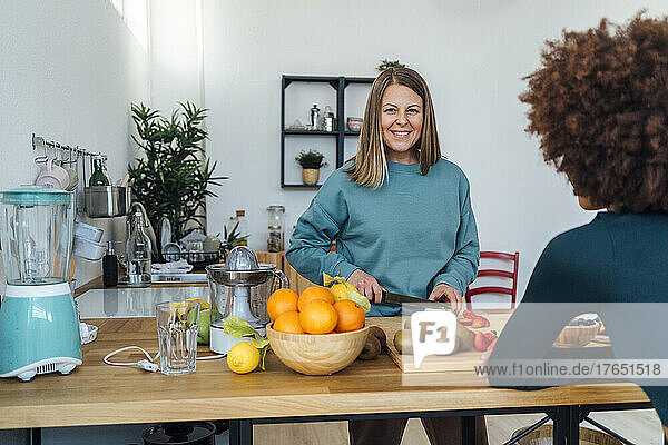 Smiling blond woman cutting apples with friend sitting at table in kitchen