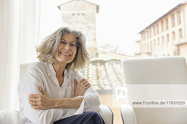 Happy senior woman with arms crossed sitting on armchair at home
