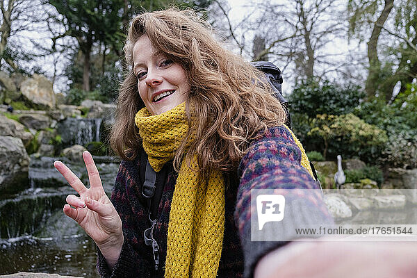 Smiling young woman gesturing peace sign taking selfie in forest