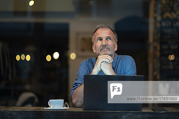 Thoughtful businessman with laptop and coffee cup sitting at table in cafe