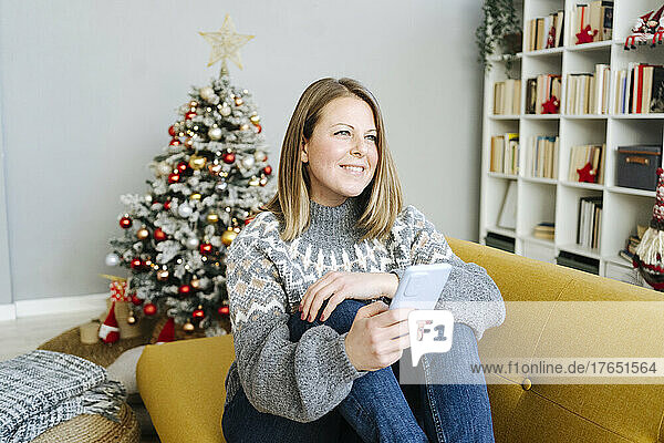 Smiling blond woman with mobile phone sitting on sofa in living room at home