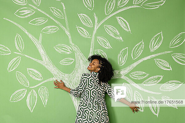 Lächelnde Frau mit geschlossenen Augen umarmt einen Baum  der eine grüne Wand zeichnet