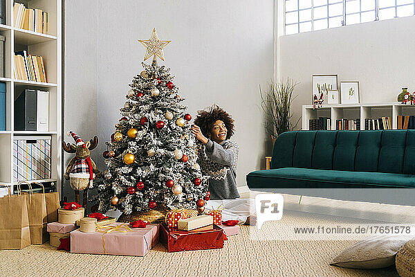 Smiling young woman sitting cross-legged decorating Christmas tree at home in living room
