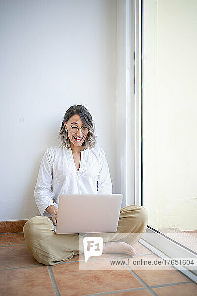 Fröhlicher junger Freiberufler  der Videoanrufe über einen Laptop führt und mit gekreuzten Beinen vor einem Glasfenster sitzt