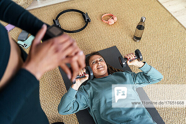 Lächelnde Frau trainiert mit Hanteln und schaut ihre Freundin im Wohnzimmer an