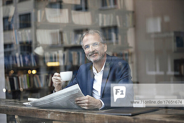 Smiling businessman with coffee cup and newspaper sitting at table in cafe