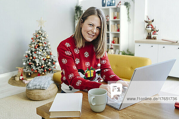 Smiling blond woman with laptop sitting at table in living room