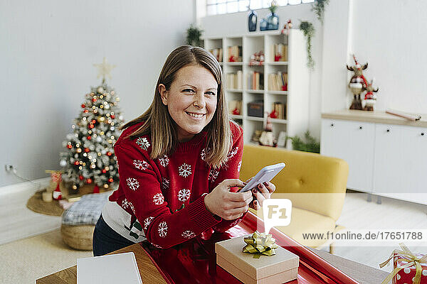 Smiling blond woman with mobile phone leaning on table at home