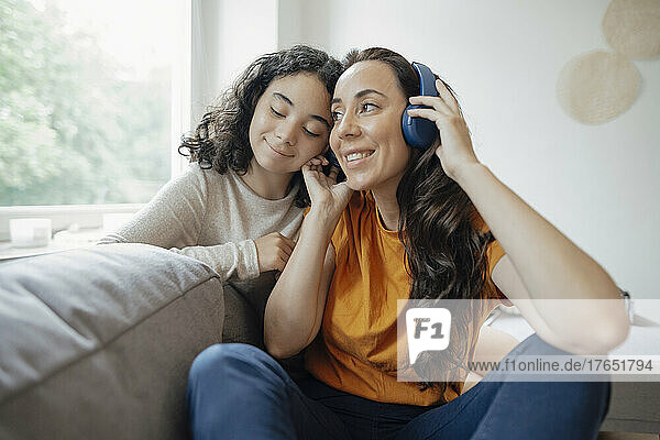 Happy woman listening music through headphones sitting with daughter on sofa at home