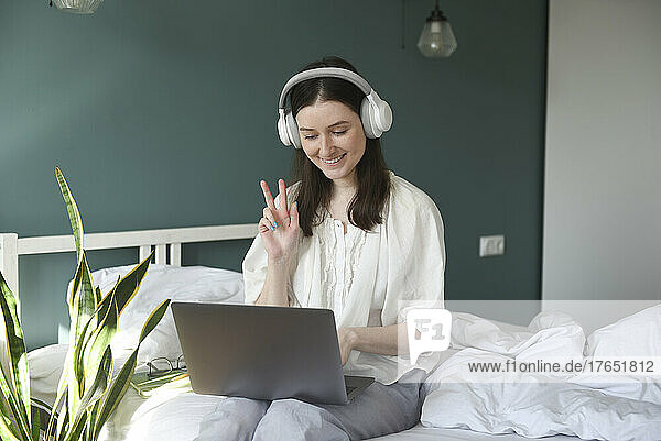 Happy woman wearing wireless headphones gesturing peace sign sitting with laptop on bed