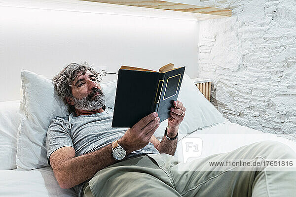 Senior man reading book lying on bed at home