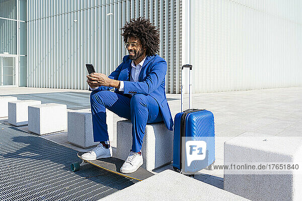 Businessman with suitcase and skateboard using mobile phone sitting on concrete block