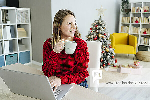 Smiling woman with coffee cup and laptop sitting at table in living room