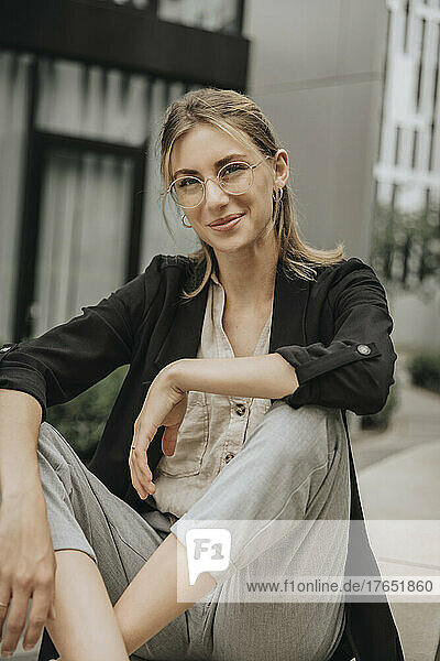 Smiling young woman wearing eyeglasses sitting on ledge
