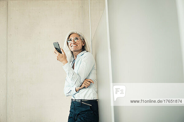 Smiling businesswoman with smart phone standing in front of wall at work place