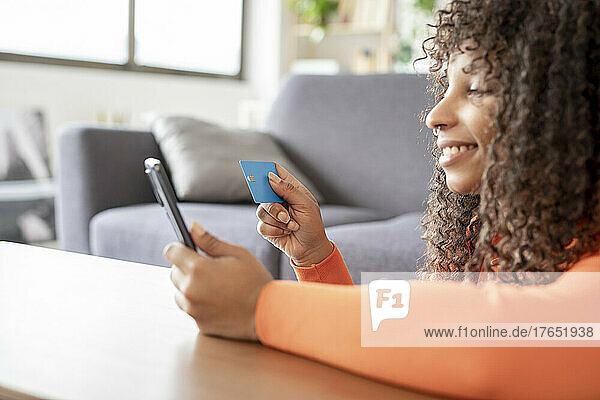 Smiling young woman with credit card using smart phone at home
