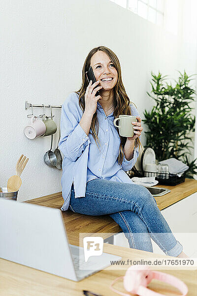 Smiling freelancer sitting on kitchen counter and talking on mobile phone at home