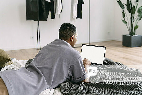 Young man using laptop lying on bedding at home