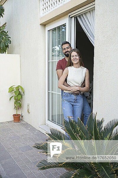 Happy couple standing by sliding door with arm around