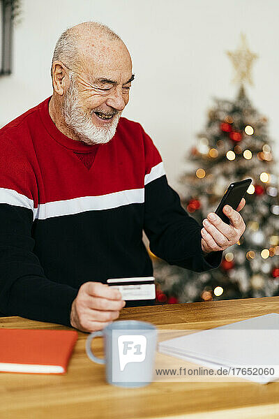 Happy senior man holding credit card doing online shopping through mobile phone at home