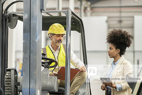 Engineer sitting in forklift discussing with businesswoman holding tablet PC in warehouse
