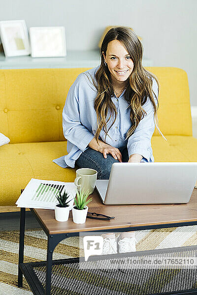 Happy freelancer with laptop sitting on yellow sofa at home