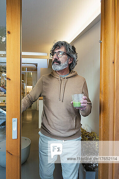 Man with glass of smoothie standing by sliding door at home