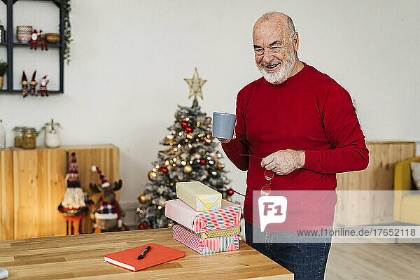 Lächelnder älterer Mann hält zu Hause eine Kaffeetasse mit Geschenkboxen auf dem Tisch