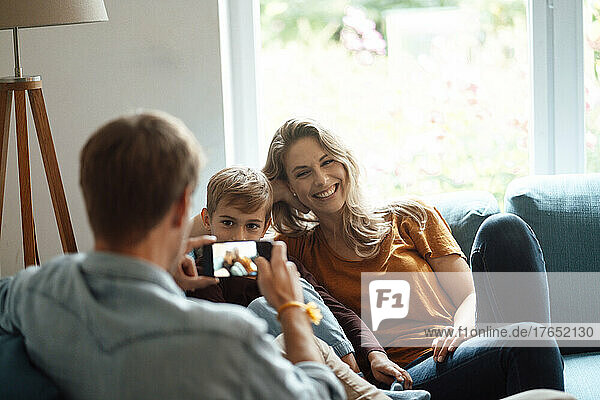 Happy blond woman and son photographed by man through smart phone in living room