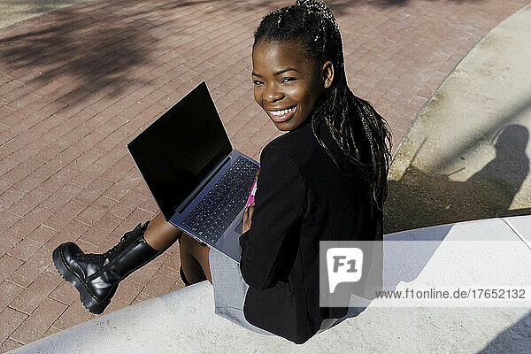Happy young woman with laptop sitting on concrete seat