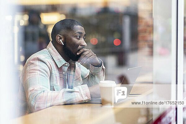 Thoughtful freelancer with laptop sitting at cafe shop seen through glass