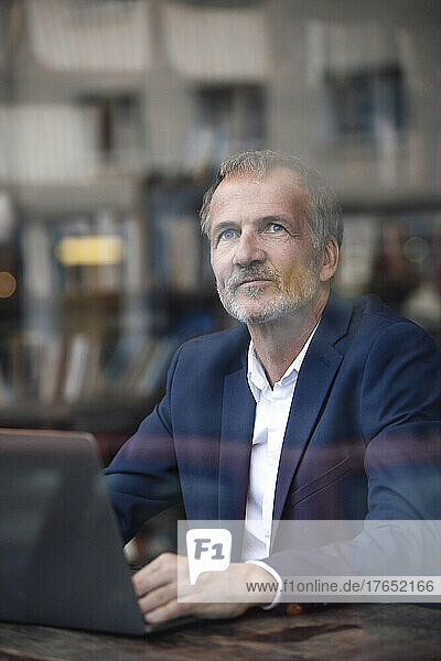 Thoughtful businessman with laptop seen through window at cafe