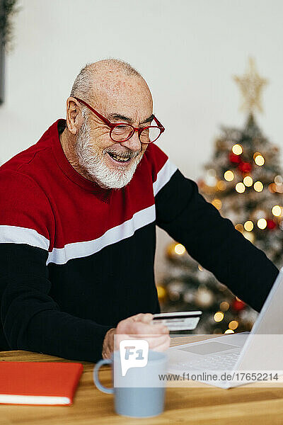 Happy senior man holding credit card doing online shopping through laptop at home
