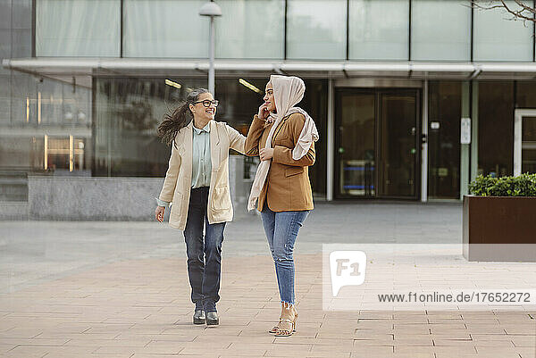 Happy businesswoman walking by young colleague standing on footpath